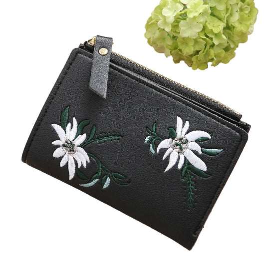 Black wallet with floral embroidery on a wooden surface with green flowers in the background